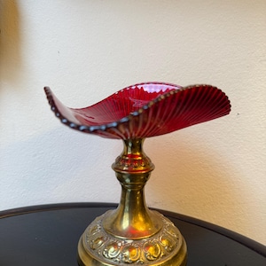 Könnte beinhalten: Eine rubinrote Glasschale mit gewelltem Rand steht auf einem Messing-Sockel. Der Sockel weist verzierte Details und ein ausgestelltes Design auf. Die Schale hat eine geschwungene, offene Form, und der Messingfuß verleiht eine Vintage-Ästhetik.