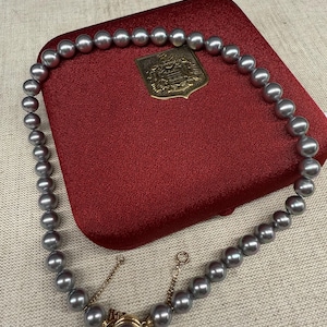 May include: A necklace of grey pearls lies on a red velvet jewellery box. The necklace is fastened with a gold clasp and chain. A gold emblem is also visible on the box.