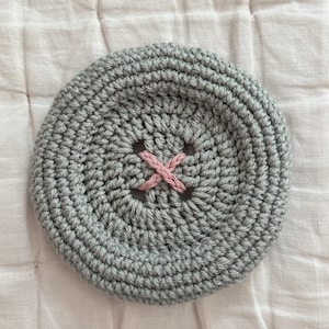 May include: A circular, grey crocheted button featuring a textured, yarn-based construction. The center of the button displays a distinct, cross-shaped stitch pattern executed in a contrasting light pink yarn, mimicking the appearance of a four-hole button.