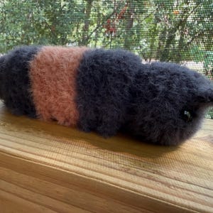 May include: A handmade plush toy in the shape of a caterpillar. The caterpillar has a fuzzy texture and is primarily dark gray with a peach-colored section in the middle. The toy is resting on a wooden surface.