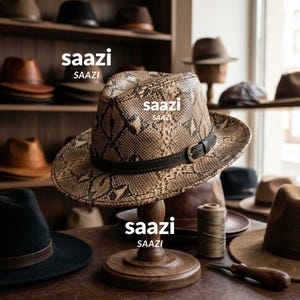 May include: A fedora hat with a tan and black snakeskin pattern, featuring a dark brown leather band with a metal buckle. The word saazi is printed on the hat and the wooden stand. The hat rests on a wooden display stand on a table.