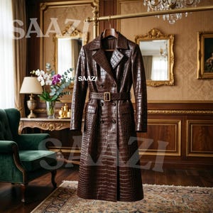 May include: A dark brown trench coat featuring a textured crocodile skin pattern, a double-breasted front with dark buttons, a matching waist belt with a gold-toned buckle, wide lapels, and long sleeves, with the text SAAZI visible on the garment.