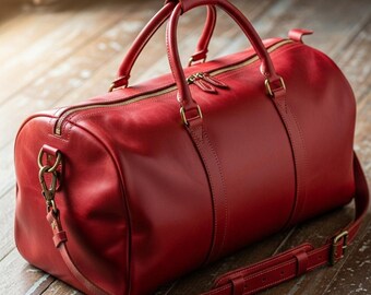 Könnte beinhalten: Eine rote Leder-Reisetasche mit einem oberen Griff und einem abnehmbaren Schultergurt. Die Tasche hat einen Reißverschluss und goldfarbene Hardware. Die Tasche steht auf einer Holzoberfläche.