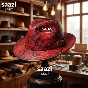 May include: A red fedora hat with a black snakeskin pattern, featuring a matching red hat band with a metal buckle. The word saazi is printed on the crown of the hat. The hat is displayed on a dark stand in a workshop setting.