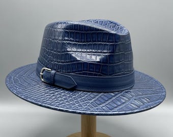 Op de afbeelding: Een blauwe fedora hoed met een krokodillenleer textuur. De hoed heeft een bijpassende blauwe band met een zilveren gesp. De hoed wordt gepresenteerd op een houten standaard tegen een neutrale achtergrond.