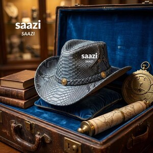 Pode incluir: Um chapéu de cowboy com textura de pele de cobra cinzenta, com uma faixa trançada e detalhes circulares dourados. O chapéu está exposto dentro de um baú vintage aberto forrado a veludo azul, junto a livros, um pergaminho e um astrolábio.