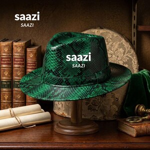 Può includere: Un cappello fedora con un vivace motivo a pelle di serpente verde e nero e una fascia abbinata. La parola saazi è stampata in bianco sulla parte anteriore della corona. Il cappello è esposto su un supporto in legno, circondato da libri antichi.