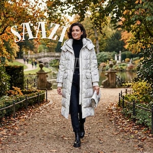 May include: A woman in a long, white and grey patterned puffer coat, black trousers, and black boots walks along a path in a park. The coat has a hood and pockets. The word "SAAZI" is displayed in large, white letters in the upper left corner.