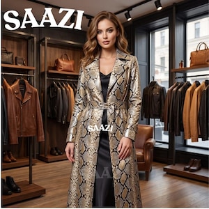 Könnte beinhalten: Ein langer, gegürteter Trenchcoat mit einem metallisch goldenen und schwarzen Schlangenledermuster, getragen über einem schlichten schwarzen Slip-Kleid. Das Wort SAAZI ist oben links und in der Mitte des Bildes in Weiß gedruckt, im Hintergrund ist ein Ladenlokal zu sehen.