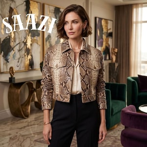 May include: A woman wearing a snakeskin-patterned jacket over a cream-colored blouse and black pants. The jacket has two chest pockets and a button-down front. The word "SAAZI" is displayed in white, stylized text. The background includes abstract art and green and purple armchairs.