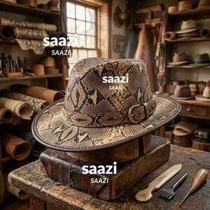 May include: A fedora hat featuring a tan and black snakeskin pattern, accented with a matching snakeskin band and a brass-toned metal buckle. The word saazi is printed on the hat and the wooden block base in white text.