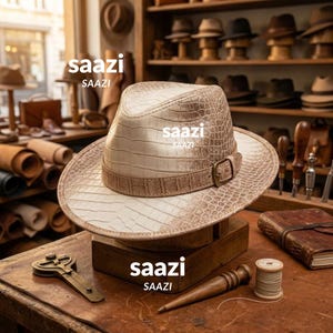 Op de afbeelding: Een beige fedora hoed met een gestructureerd krokodillenleerpatroon, voorzien van een bijpassende band met een metalen gesp. Het woord saazi staat op de hoed en de houten voet gedrukt. De hoed staat op een houten standaard in een werkplaats.