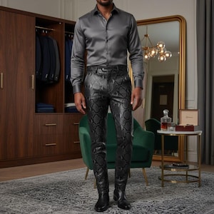 May include: A man wearing a gray satin button-down shirt and black snake-skin patterned pants. The pants are fitted and have a dark gray and black pattern. The man is wearing black boots. A wooden wardrobe is in the background.