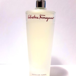 Könnte beinhalten: Ein rechteckiger Parfümflakon mit silbernem Verschluss und mattiertem Glas. Der Schriftzug "Salvatore Ferragamo" in Rot und "TUSCAN SOUL" sind auf der Vorderseite aufgedruckt. Der Flakon enthält eine hellgelbe Flüssigkeit.