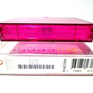 May include: A pink perfume bottle and its packaging. The box is white with the words "GUCCI rush" in pink and orange. The perfume bottle is a bright pink colour. The set includes a barcode and other product information.