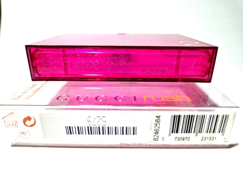 May include: A pink perfume bottle and its packaging. The box is white with the words "GUCCI rush" in pink and orange. The perfume bottle is a bright pink colour. The set includes a barcode and other product information.