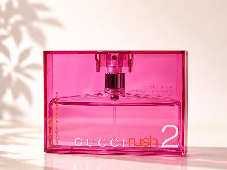 May include: A vibrant pink perfume bottle with a clear glass interior. The bottle features a silver atomizer and the words "GUCCI Rush 2" are printed in white. The words "Vaporisateur Natural Spray" are printed on the side.