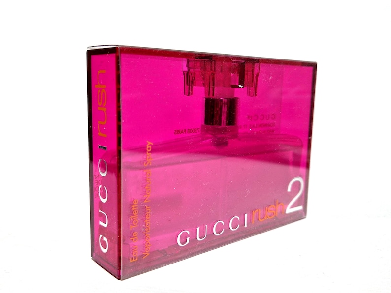May include: A rectangular, transparent pink perfume bottle with a silver atomizer. The bottle is labelled "GUCCI Rush 2" in white lettering. The words "Eau de Toilette Vaporisateur Natural Spray" are also visible.