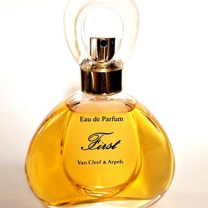 Può includere: Un flacone di profumo con un tappo trasparente a forma di anello e un vaporizzatore color oro. Il flacone è riempito con un liquido color oro e presenta le scritte "Eau de Parfum" e "First Van Cleef & Arpels".