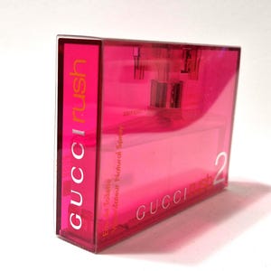 May include: A rectangular, transparent pink perfume bottle with the text "GUCCI rush" in orange and white. The bottle also has the text "Eau de Toilette Vaporisateur Natural Spray" and the number "2".