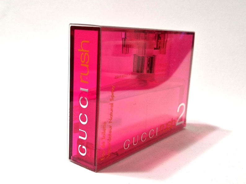 May include: A rectangular, transparent pink perfume bottle with the text "GUCCI rush" in orange and white. The bottle also has the text "Eau de Toilette Vaporisateur Natural Spray" and the number "2".