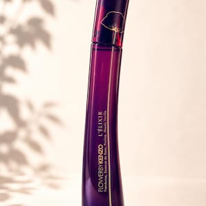 Könnte beinhalten: Eine violette Parfümflasche mit goldenem Blumendesign und dem Text "FLOWERYBYKENZO L'ÉLIXIR". Die Flasche hat eine geschwungene Form und einen dunkelvioletten Deckel. Der Hintergrund ist eine weiche, neutrale Farbe mit Blätterschatten.