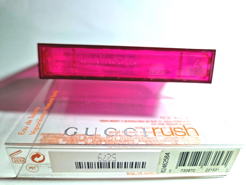 May include: A vibrant pink perfume bottle and box set. The box displays the brand name "Gucci Rush". The transparent bottle sits above the box, which has a barcode and other printed details.
