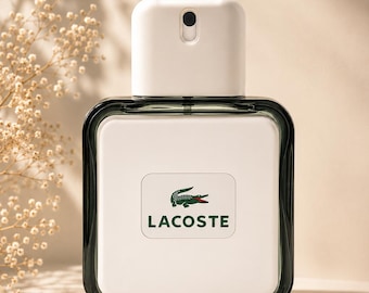 May include: A rectangular perfume bottle with a white cap and a dark green border. The bottle features the Lacoste logo, a green crocodile, and the brand name in green. The background is a soft beige with subtle shadows.