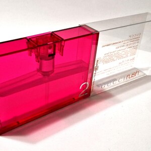 May include: A rectangular, bright pink perfume bottle in a clear plastic case. The bottle has a spray nozzle and the number "2" is visible. The clear case has the words "Gucci Rush" and other text.
