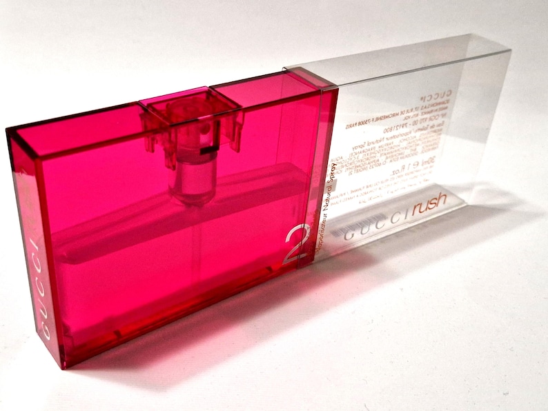 May include: A rectangular, bright pink perfume bottle in a clear plastic case. The bottle has a spray nozzle and the number "2" is visible. The clear case has the words "Gucci Rush" and other text.