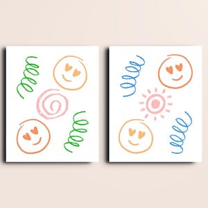 May include: Two white art prints with colourful doodles. Each print features orange smiley faces with heart eyes, green and blue squiggles, a pink spiral, and a pink sun.