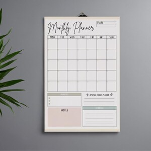 Peut inclure: Un planificateur mensuel rectangulaire avec une bordure crème, accroché à un mur gris. Le planificateur comprend une grille pour les jours du mois, avec des sections pour les priorités, les notes et les listes de tâches. Le texte "Monthly Planner" est écrit en haut.