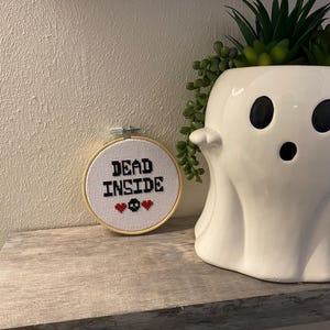 May include: A white ceramic ghost-shaped planter with a succulent plant inside. Next to it is a cross-stitch hoop with the words "DEAD INSIDE" and skull and heart symbols. The items are on a wooden shelf.