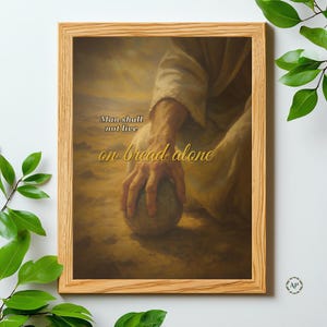 Op de afbeelding: Een ingelijste prent met een religieuze scène met een hand die een steen vasthoudt. De tekst "Man shall not live on bread alone" staat in gouden letters. Het kunstwerk heeft een warme, aardse achtergrond.