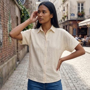 Puede incluir: Una camisa amarilla clara de manga corta, abotonada y con cuello clásico. La camisa se lleva con vaqueros azules. El escenario es una calle adoquinada.