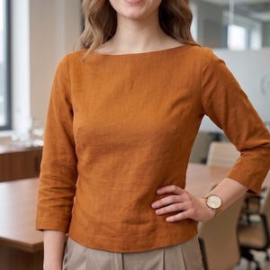 May include: A burnt orange, boat-neck top with three-quarter length sleeves. The top is made of a textured fabric and has a relaxed fit. The person is wearing a gold watch. The top is a warm, earthy tone.