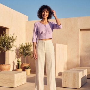 May include: A light purple, textured, cropped button-down top with three-quarter length sleeves and a round neckline, paired with high-waisted, wide-leg cream trousers featuring a clean, tailored silhouette and a central vertical seam on each leg.