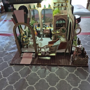 Może przedstawiać: Miniaturowy drewniany model salonu fryzjerskiego z szczegółowym wnętrzem. Zawiera lustra, krzesła i narzędzia do stylizacji. Znak nad wejściem głosi "hair salon". Model umieszczony jest na drewnianej podłodze z różowym dywanem.