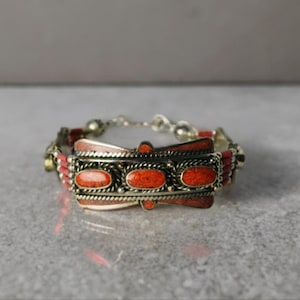 Könnte beinhalten: Silberfarbenes Armband mit einem zentralen rechteckigen Design mit drei ovalen, orangeroten Steinen. Das Armband hat einen Kettenverschluss und kleinere rote Perlen, die einen Vintage- oder Ethno-Schmuckstil erzeugen.