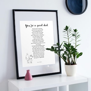 May include: A black framed print featuring the text You're a good dad followed by a poem and a line drawing of an adult holding a child's hand. The print is white with black lettering and sits on a white surface next to a green plant.