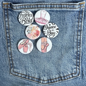 May include: A collection of round, white pinback buttons with ballet-themed designs. One button reads "Be Fearless Be Bold." Other buttons feature a pink tutu, ballet shoes, and the text "Dance All Day." The buttons are arranged on a denim background.