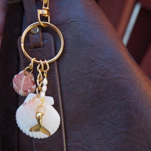 May include: A gold-toned metal keychain featuring a circular ring, a small pink and white scallop shell, a larger white scallop shell, a small chain with white pearls, and a gold-toned metal whale tail charm attached to a dark brown leather bag.