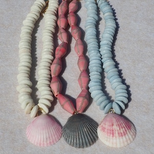 May include: Three necklaces featuring strands of beads and shell pendants. The left necklace has white disc beads and a pink shell. The centre necklace has pink spindle-shaped beads with gold lines and a grey shell. The right has light blue disc beads and a pink and white shell.