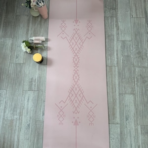 Può includere: Un tappetino da yoga rosa chiaro con un motivo geometrico centrale in una tonalità di rosa più chiara. Il tappetino è rettangolare e presenta una linea verticale centrale. Sono visibili altri oggetti, tra cui un bicchiere, fiori e prodotti per la cura della pelle.