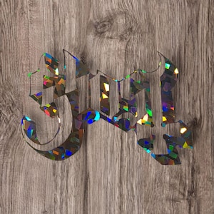 May include: Holographic "Ghost" logo sticker with a gothic font style. The sticker has a rainbow iridescent finish and is set against a wood grain background. The word "Ghost" is displayed.