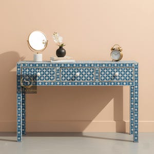 Puede incluir: Una mesa de tocador con estampado azul y blanco con tres cajones y pomos blancos. Un espejo con marco dorado, un pequeño jarrón negro con una flor blanca y un despertador dorado encima. La mesa tiene un diseño geométrico.