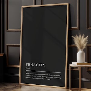 May include: A framed black print featuring the word "TENACITY" in white, accompanied by a definition. The print is propped against a dark wall in a room with a window and a small side table with a vase and decorative plants.
