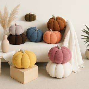May include: A collection of plush pumpkin-shaped pillows in various colors, including orange, yellow, pink, blue, and brown. Each pillow has a stem and is arranged on a white sofa and a wooden block, creating a cozy, autumnal theme.