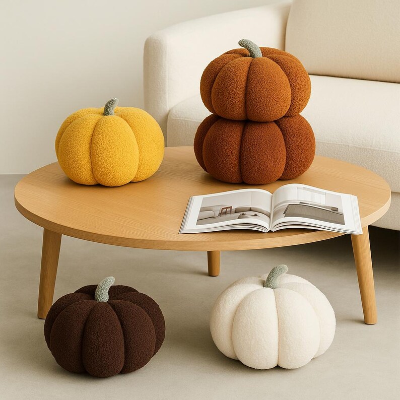 May include: A collection of plush pumpkin-shaped cushions in various colors, including yellow, orange, brown, and white. The pumpkins have a textured, fuzzy appearance and green stems. They are arranged on a wooden coffee table with an open book.