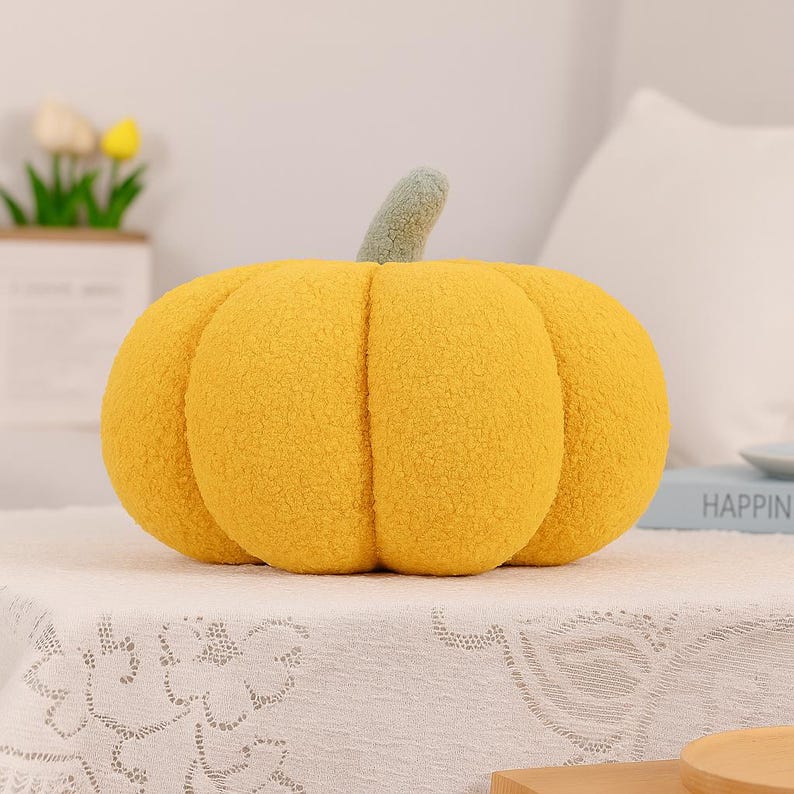 May include: A plush, yellow pumpkin-shaped pillow with a green stem. The pillow has a textured surface and is sitting on a white tablecloth. The pumpkin is a decorative item, perfect for fall or Halloween.